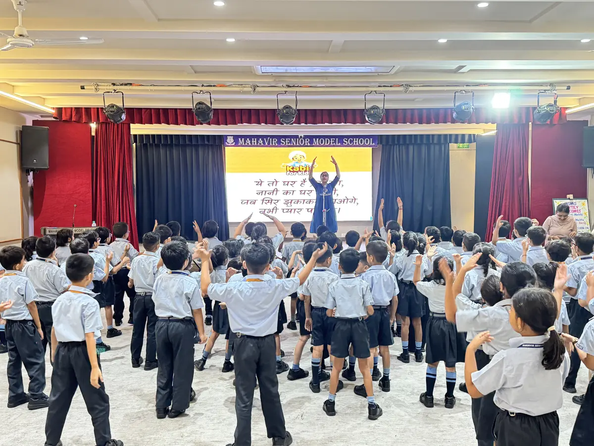 The whole of Class 2 on their feet in the hall at Mahavir Senior Model School, arms raised, learning the steps to a Little Kabir Song