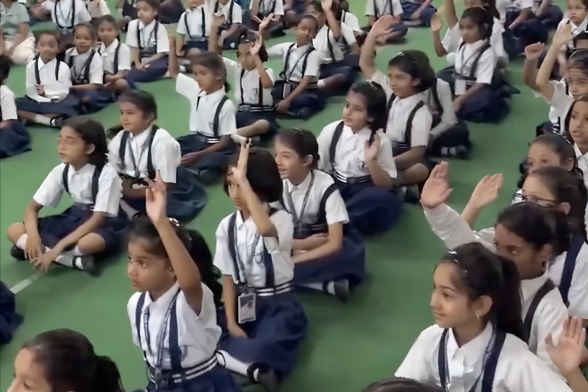 Young girls at St Mary's Convent Junior, Allahabad, seated on a green-floored hall with hands raised during a K4K session, April 2026